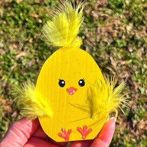 May include: A wooden, egg-shaped chick decoration painted bright yellow. It features black eyes, a pink beak, and small pink feet. Yellow feathers adorn the top and sides, creating a whimsical Easter-themed ornament.