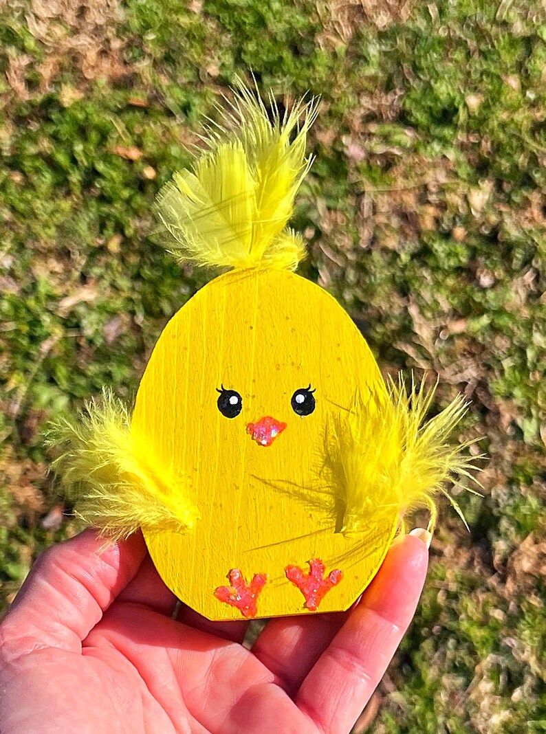 May include: A wooden, egg-shaped chick decoration painted bright yellow. It features black eyes, a pink beak, and small pink feet. Yellow feathers adorn the top and sides, creating a whimsical Easter-themed ornament.