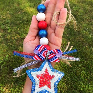 May include: A red, white and blue beaded garland with a star-shaped ornament. The star is decorated with red, white and blue glitter and has a red star in the centre. The garland is tied with a red, white and blue ribbon.