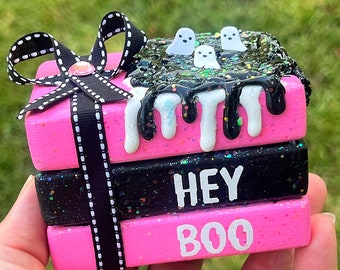 May include: A stack of three pink and black wooden blocks with glitter. The top block has white dripping paint and three white ghost figures. The middle block says "HEY" in white letters. The bottom block says "BOO" in white letters. A black and white ribbon with a rhinestone is tied around the stack.