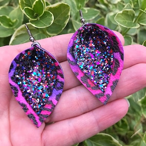 May include: A pair of dangle earrings with a pink, purple, and black leopard print design. The earrings have a silver hook and a clear resin center filled with colorful glitter.