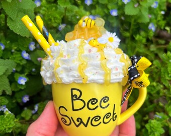 May include: A yellow ceramic mug with a white whipped cream topping, honey drizzle, and bee decorations. The mug has the text "Bee Sweet" written on it.