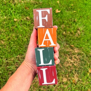 May include: Four wooden blocks stacked vertically spelling out the word "FALL". The blocks are painted in different colors: brown, orange, green, and red. Each letter is painted in white.