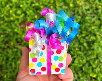May include: A small gift box with a white background and colorful polka dots. The box is wrapped with pink, blue, and white ribbon.