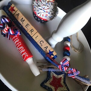 May include: A white tiered serving tray decorated with red, white and blue. The tray features a cupcake with red, white and blue frosting, a wooden sign that reads "AMERICA", a red and white rolling pin with the word "FIREWORKS" written on it, a blue and white star with glitter, and a blue and white beaded garland.
