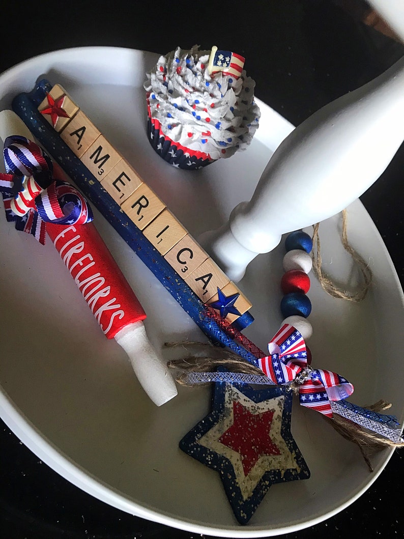 May include: A white tiered serving tray decorated with red, white and blue. The tray features a cupcake with red, white and blue frosting, a wooden sign that reads "AMERICA", a red and white rolling pin with the word "FIREWORKS" written on it, a blue and white star with glitter, and a blue and white beaded garland.