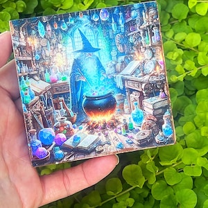 May include: A square wooden tile featuring a detailed illustration of a wizard in a blue robe and hat, stirring a bubbling cauldron. The scene is filled with potion bottles, books, and magical artifacts, with a mystical, glowing effect.