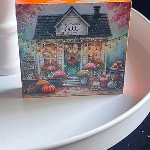May include: A decorative wooden block featuring a fall-themed shop scene. The image shows a shop with pumpkins, flowers, and the text "ALL THINGS fall". The block has an orange and blue top, and is set on a white surface.