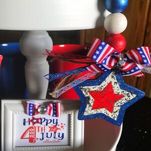 May include: A white tiered tray decorated for the 4th of July with red, white and blue accents. The tray features a small framed sign that says "Happy 4th of July" with a red, white and blue bow on top. There is also a red, white and blue star with glitter on top of the tray.