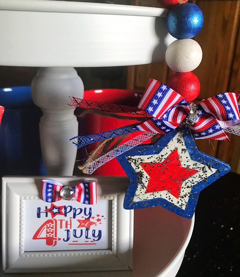 May include: A white tiered tray decorated for the 4th of July with red, white and blue accents. The tray features a small framed sign that says "Happy 4th of July" with a red, white and blue bow on top. There is also a red, white and blue star with glitter on top of the tray.