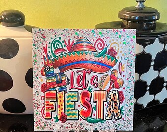 May include: A white square sign with glitter and colorful images of a sombrero, a pinata, maracas, and chili peppers. The text "Let's Fiesta" is in large, colorful letters.