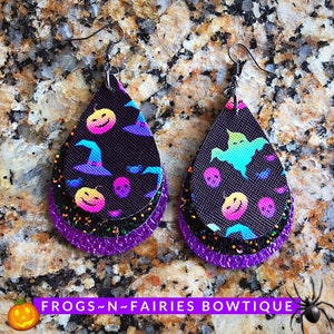 May include: A pair of layered teardrop-shaped earrings with a black background and a Halloween theme. The earrings feature a variety of colorful Halloween icons, including pumpkins, ghosts, witches' hats, and skulls. The earrings are accented with purple glitter.