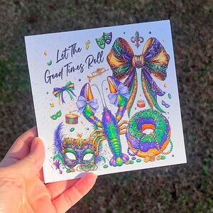Pode incluir: Cart&atilde;o quadrado com uma ilustra&ccedil;&atilde;o colorida apresentando uma lagosta, uma m&aacute;scara, um la&ccedil;o e uma rosquinha, com o texto "Let The Good Times Roll". O design incorpora elementos roxos, verdes, dourados e azuis, evocando um tema festivo.
