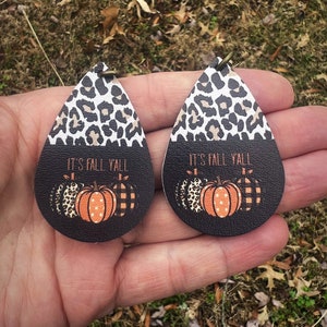 May include: A pair of dangle earrings with a black and white leopard print design. The earrings feature a black background with the text "It's Fall Y'all" and three orange pumpkins with white polka dots, black and white leopard print, and black and white checkered patterns.