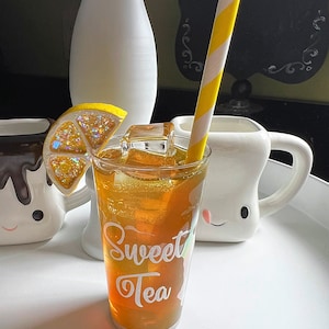 May include: A glass of iced sweet tea with a lemon wedge and ice cubes. The glass has the words "Sweet Tea" printed on it. A yellow and white striped straw is in the glass.