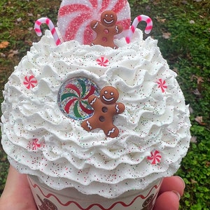 May include: A white cupcake topped with red and white candy canes, a red and white peppermint candy, and two gingerbread men. The cupcake is decorated with white frosting and red and white sprinkles. The cupcake is in a white paper liner with the words "Warm Wishes" printed on the side.