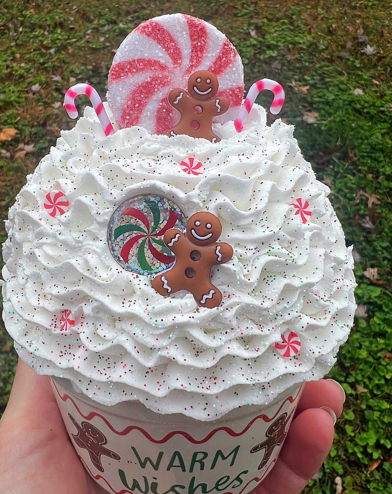 May include: A white cupcake topped with red and white candy canes, a red and white peppermint candy, and two gingerbread men. The cupcake is decorated with white frosting and red and white sprinkles. The cupcake is in a white paper liner with the words "Warm Wishes" printed on the side.