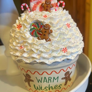 May include: White cupcake decorated with red and white candy canes, red and white peppermint candies, and gingerbread men. The cupcake is topped with white frosting and glitter. The cupcake is in a white cup with red and green trim and the words "Warm Wishes" printed on the front.