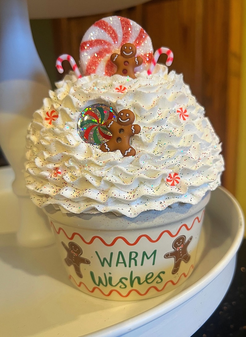 May include: White cupcake decorated with red and white candy canes, red and white peppermint candies, and gingerbread men. The cupcake is topped with white frosting and glitter. The cupcake is in a white cup with red and green trim and the words "Warm Wishes" printed on the front.