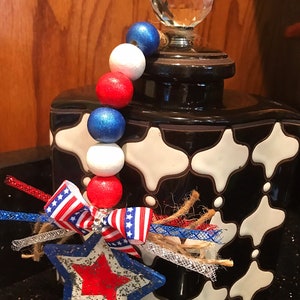 May include: A black and white ceramic jar with a crystal knob on the lid. The jar is decorated with a red, white and blue beaded garland and a star-shaped ornament with a red, white and blue ribbon.