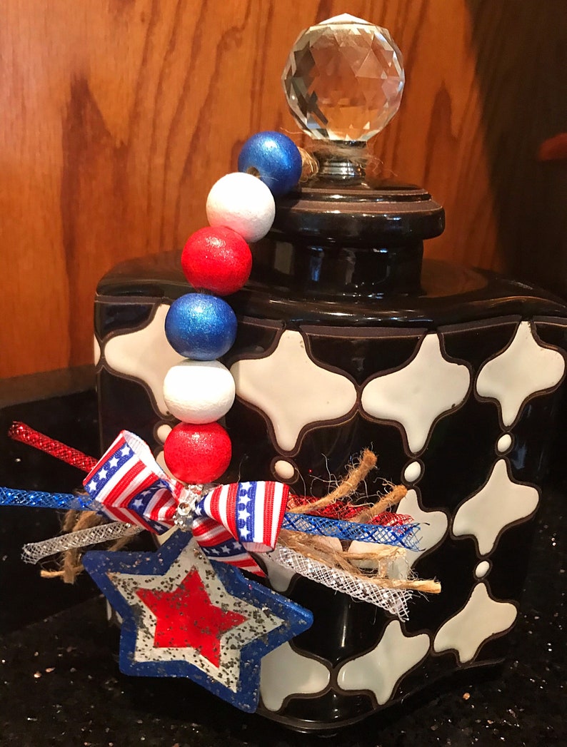 May include: A black and white ceramic jar with a crystal knob on the lid. The jar is decorated with a red, white and blue beaded garland and a star-shaped ornament with a red, white and blue ribbon.