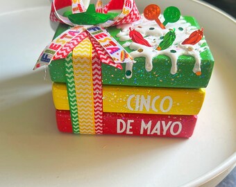 May include: A stack of three small wooden boxes decorated for Cinco de Mayo. The top box is green with white frosting and red and green peppers. The middle box is yellow with the word "CINCO" in white letters. The bottom box is red with the words "DE MAYO" in white letters. The boxes are tied together with a red and yellow ribbon.