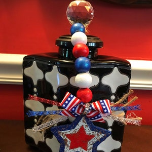 May include: Black and white ceramic jar with a red, white and blue star and ribbon decoration. The jar has a crystal knob on the lid and a beaded garland hanging from the knob.