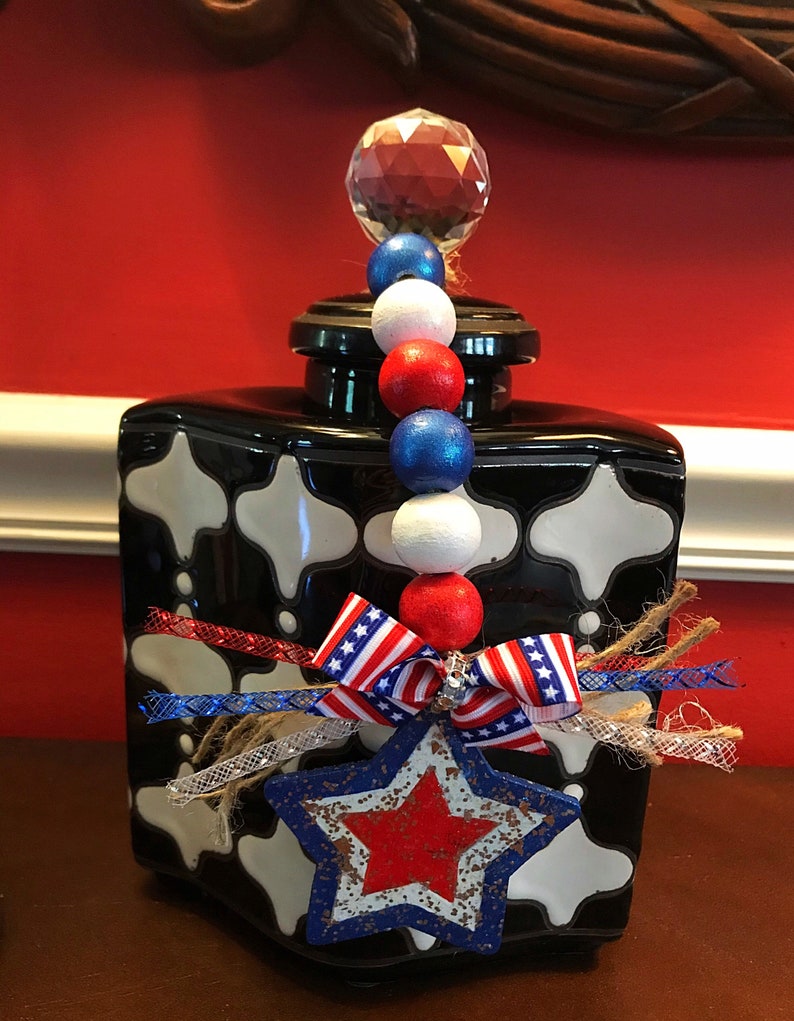 May include: Black and white ceramic jar with a red, white and blue star and ribbon decoration. The jar has a crystal knob on the lid and a beaded garland hanging from the knob.