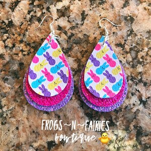 May include: Teardrop-shaped earrings with a layered design. The top layer features a white background with colorful bunny prints. The middle layer is pink, and the bottom layer is purple glitter. Silver hooks complete the earrings. The text "FROGS ~N~ FAIRIES boutique" is at the bottom.