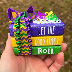 May include: A stack of three wooden blocks decorated with purple, yellow, and green paint. The blocks have the words "Let the good times roll" written on them. The blocks are decorated with a purple ribbon and green and purple beads.