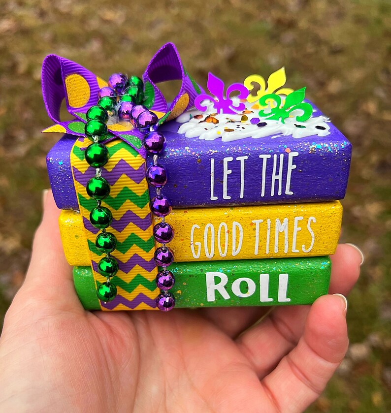 May include: A stack of three wooden blocks decorated with purple, yellow, and green paint. The blocks have the words "Let the good times roll" written on them. The blocks are decorated with a purple ribbon and green and purple beads.