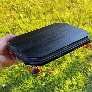 May include: A black wooden serving tray with an octagonal shape and distressed edges. The tray is supported by four amber-colored glass knobs. The tray is held over a background of green grass.