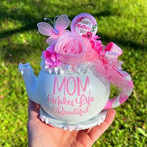 May include: White teapot with a pink and white floral arrangement, a pink butterfly, and the text "MOM Makes Life Beautiful". The teapot has a white saucer and a pink polka dot bow.