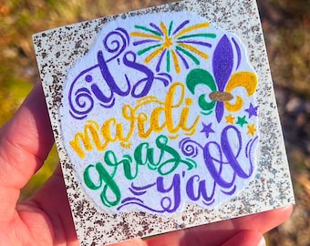 May include: A white square sign with glitter on the edges. The sign has a purple, green, and gold design with the text "It's Mardi Gras y'all".