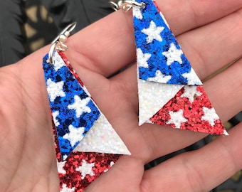 May include: A pair of dangle earrings with a red, white, and blue glitter design. The earrings are shaped like triangles and feature white stars on each color.