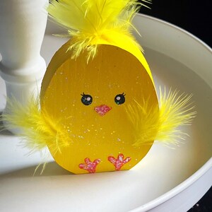 May include: A yellow wooden Easter chick decoration with yellow feather accents. The chick has painted eyes, a pink beak, and pink feet. The decoration is on a white tray, perfect for Easter decor.