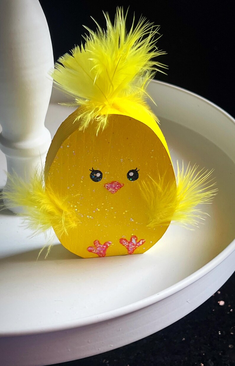 May include: A yellow wooden Easter chick decoration with yellow feather accents. The chick has painted eyes, a pink beak, and pink feet. The decoration is on a white tray, perfect for Easter decor.