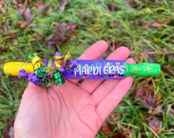 Puede incluir: Un silbato de Mardi Gras morado y verde con purpurina dorada y serpentinas amarillas y verdes.