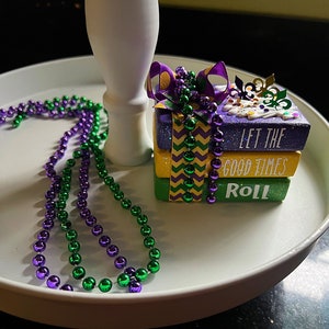 May include: A tiered tray decorated with purple and green Mardi Gras beads and a stack of three purple, green, and gold gift boxes with the text "Let the good times roll".