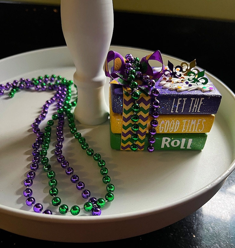 May include: A tiered tray decorated with purple and green Mardi Gras beads and a stack of three purple, green, and gold gift boxes with the text "Let the good times roll".