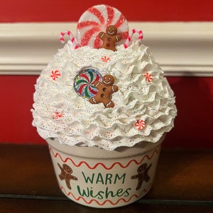 May include: White ceramic cupcake container with red and white candy cane accents, a red and white peppermint candy, and gingerbread men. The container has the words "Warm Wishes" written on it.