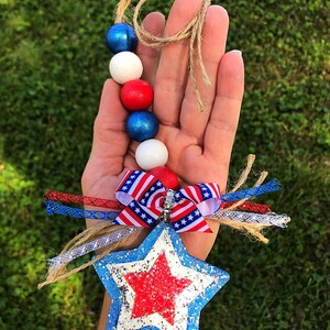 May include: A red, white and blue beaded garland with a star-shaped ornament. The star is blue with a red glitter star in the centre. The garland is tied with a red, white and blue ribbon.