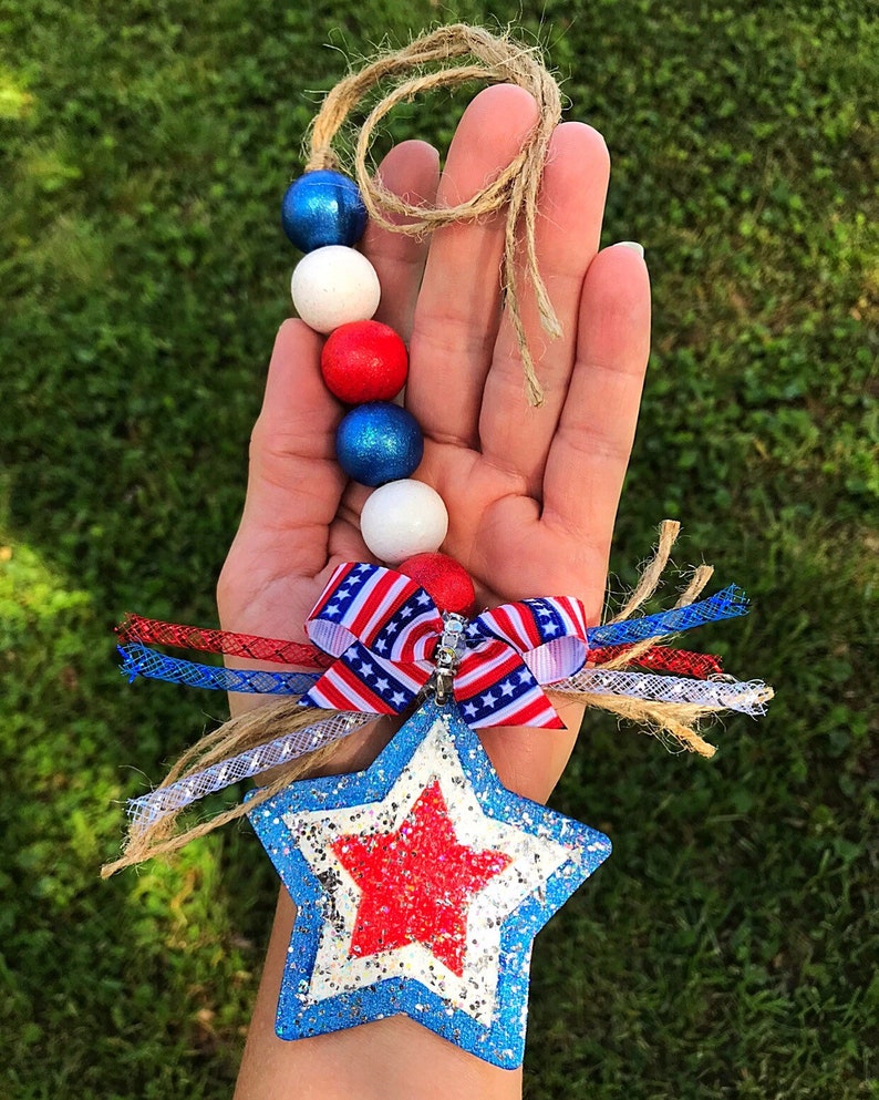 May include: A red, white and blue beaded garland with a star-shaped ornament. The star is blue with a red glitter star in the centre. The garland is tied with a red, white and blue ribbon.