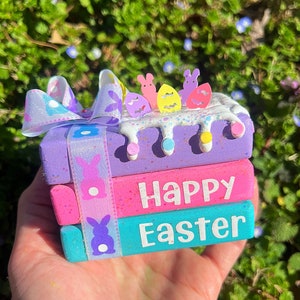 May include: A stack of three wooden blocks decorated with pastel colors, glitter, and bunny and egg shapes. The top block has a white background with pink, yellow, and blue accents. The middle block is pink with white polka dots and the text "Happy" in white. The bottom block is turquoise with white polka dots and the text "Easter" in white.
