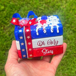 May include: A stack of three wooden blocks decorated with red, white, and blue stars and glitter. The top block has a white background with blue and white glitter and a red, white, and blue ribbon. The middle block has a white background with the text "Oh My" in black. The bottom block has a red background with the text "Stars" in white.