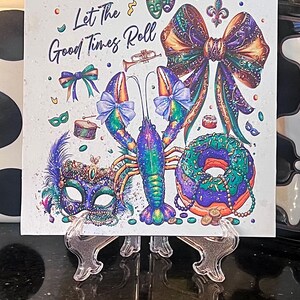 Pode incluir: Azulejo quadrado com um design colorido de Mardi Gras. Apresenta uma lagosta, um la&ccedil;o, uma m&aacute;scara e uma rosquinha com o texto "Let The Good Times Roll". A obra de arte &eacute; em tons de roxo, verde e dourado.