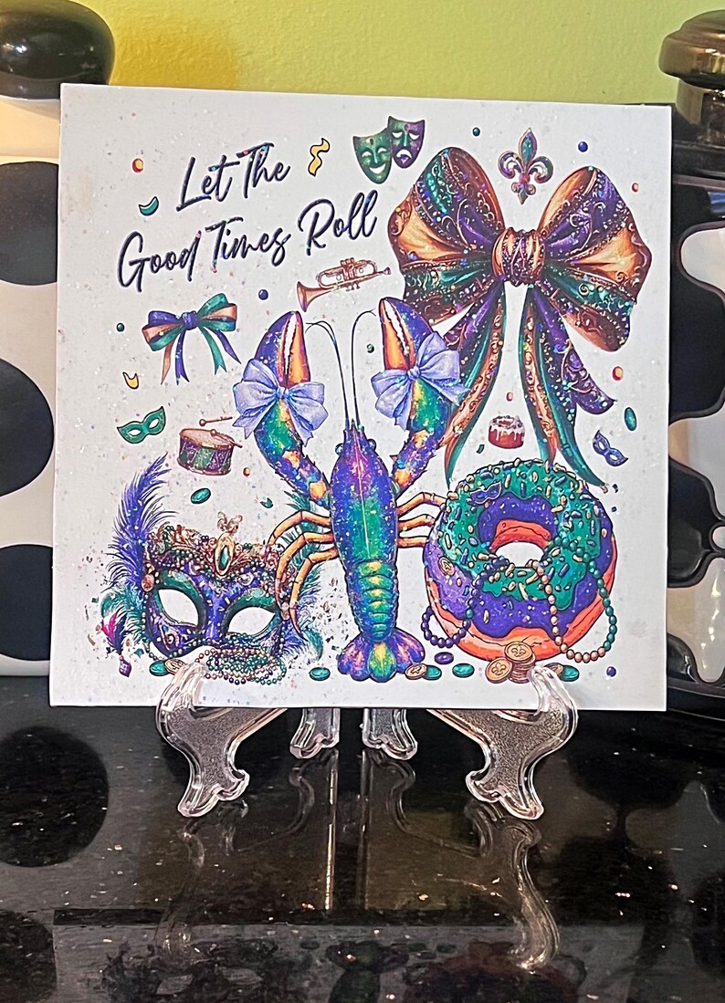 Pode incluir: Azulejo quadrado com um design colorido de Mardi Gras. Apresenta uma lagosta, um la&ccedil;o, uma m&aacute;scara e uma rosquinha com o texto "Let The Good Times Roll". A obra de arte &eacute; em tons de roxo, verde e dourado.
