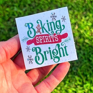 May include: A square, white decorative item with the text "Baking Spirits Bright" in green and red. The design includes a gingerbread man, snowflakes, and a rolling pin. The item is held in a hand against a green background.