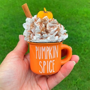 May include: Orange ceramic mug with white lettering that reads "Pumpkin Spice". The mug is topped with white whipped cream, a cinnamon stick, and a small orange pumpkin.