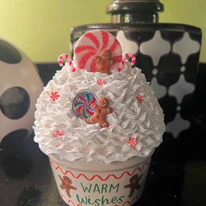 May include: A white ceramic cupcake container with a white frosting-like texture and red and white candy cane accents. The container has a red and white peppermint candy on top and two gingerbread men. The container has the words "Warm Wishes" written on the front.